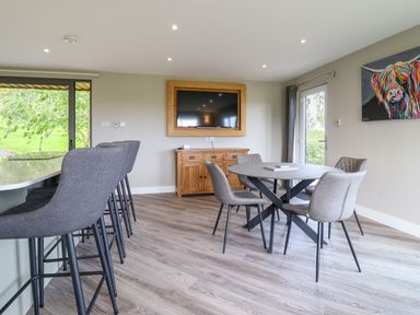 A dining area with gray chairs around a round table next to a wooden cabinet and mounted TV at Orchard Lodge in Maerdy near Corwen