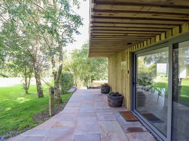 A patio with stone tiles and wooden wall next to a grassy area with trees at Orchard Lodge in Maerdy near Corwen