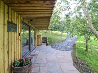 A wooden exterior patio area with stone tiles flower pot and gravel pathway surrounded by grass and trees at Orchard Lodge Maerdy near Corwen