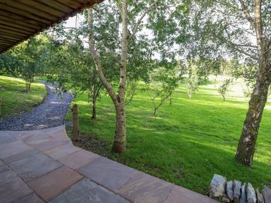 A stone patio overlooking a grassy yard with several trees and a gravel path at Orchard Lodge Maerdy near Corwen