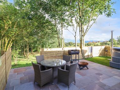 An outdoor patio with a round glass table and four chairs a barbecue a fire pit a hot tub and wooden fencing at Orchard Lodge Maerdy near Corwen