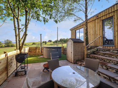 A backyard patio with a glass table and chairs a barbecue a hot tub and wooden shed beside a wooden building at Orchard Lodge in Maerdy near Corwen