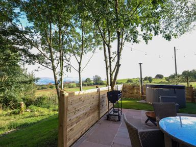 An outdoor patio with a round glass table and chairs a barbecue grill a hot tub and string lights at Orchard Lodge in Maerdy near Corwen