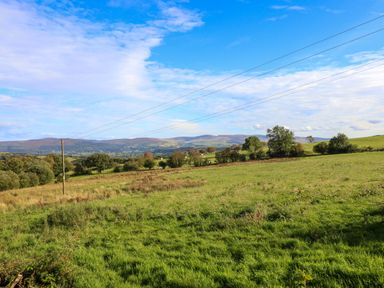 A green field with trees and hills in the background under a blue sky with clouds and power lines crossing the field at Orchard Lodge in Maerdy near Corwen