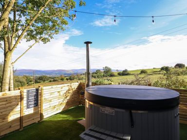 An outdoor area with a covered hot tub next to a wooden fence and a tree with string lights above at Orchard Lodge Maerdy near Corwen