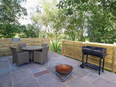 An outdoor patio with a round table and four chairs a fire pit and a grill with wooden fencing and trees at Orchard Lodge in Maerdy near Corwen