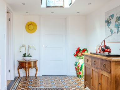 An entryway with a wooden table holding a plant a yellow hat on the wall a large floral vase with red flowers a wooden cabinet with a red helicopter model and a world map on the wall at Howard Cottage North Littleton near Offenham