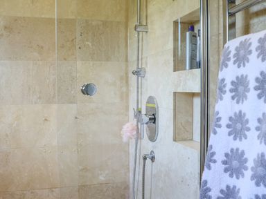 A shower area with beige tiled walls a built-in niche holding bottles a showerhead and a towel with floral patterns at Howard Cottage in North Littleton near Offenham