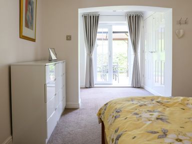 A bedroom with a white dresser a bed with yellow floral bedding glass doors and grey curtains at Howard Cottage in North Littleton near Offenham