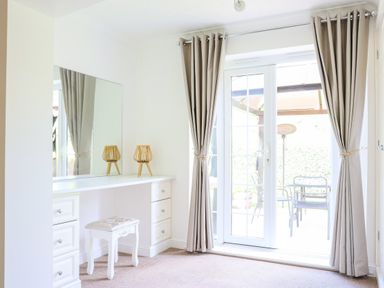 A room with a white dressing table with drawers a stool and two lamps next to glass doors with beige curtains at Howard Cottage North Littleton near Offenham