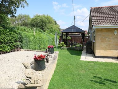A garden with green grass a gravel area flower pots a statue and a gazebo next to a house at Howard Cottage in North Littleton near Offenham