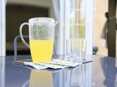 A glass pitcher filled with yellow liquid and three empty glasses on a reflective table at Howard Cottage in North Littleton near Offenham