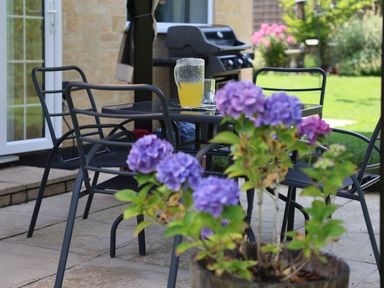 A patio with black metal chairs and table a jug of yellow drink a grill and purple flowers at Howard Cottage North Littleton near Offenham