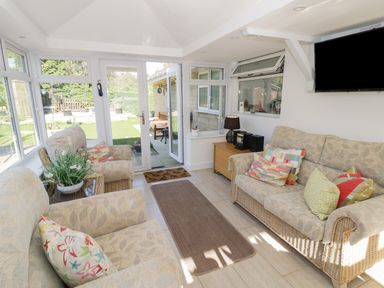 A sunroom with wicker sofas and armchairs with cushions a rug a wooden cabinet with a lamp and stereo and a wall-mounted television at Howard Cottage North Littleton near Offenham