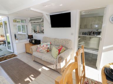 A small living area with a sofa and cushions a wooden side table with a lamp a wall-mounted TV and a view into a kitchen at Howard Cottage North Littleton near Offenham