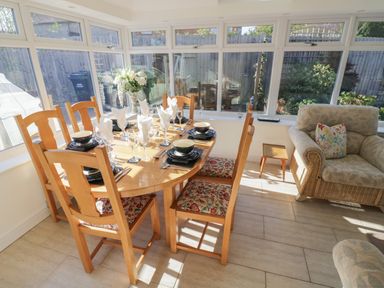 A dining area with a wooden table set with plates and glasses surrounded by wooden chairs with floral cushions and a sitting area with wicker armchairs at Howard Cottage in North Littleton near Offenham