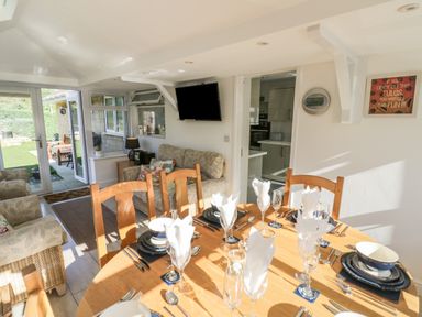 A dining area with a wooden table set with plates glasses and napkins next to a seating area with armchairs and a wall-mounted television at Howard Cottage in North Littleton near Offenham