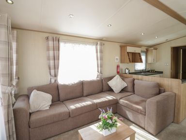 A living room with a brown sectional sofa two white pillows a small wooden coffee table with a flower arrangement beige curtains and a view of a kitchen area at Elm in Denny