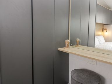 A bedroom with grey wardrobes a wooden shelf a mirror a small lamp and a round grey stool at Elm in Denny