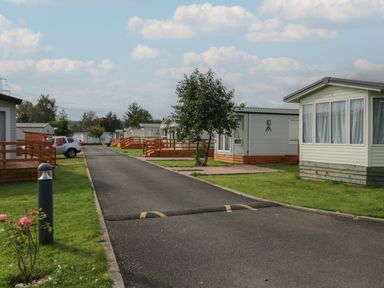 A paved road between rows of white mobile homes with small gardens and wooden fences at Elm in Denny