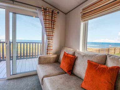A living area with a beige sofa and orange cushions next to sliding glass doors and windows overlooking a balcony and the sea at Seaview Lodge in Bacton
