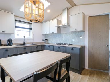 A kitchen with a wooden dining table and black chairs white and dark cabinets a window and a wooden door at Seaview Lodge in Bacton