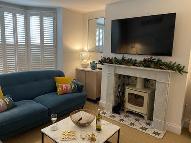 A living room with a blue sofa and colorful pillows a coffee table with wine glasses and a tic tac toe game a fireplace with greenery and a TV mounted above at Happy Place in Sheringham