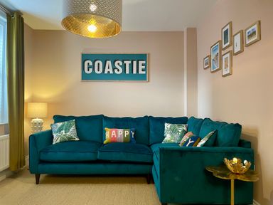 A living room with a teal sectional sofa and patterned pillows under a COASTIE wall sign at Happy Place in Sheringham