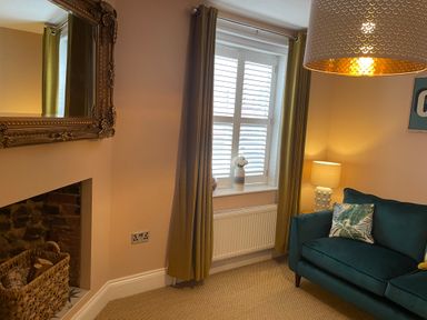 A corner of a living room with a teal sofa with a leaf patterned cushion a window with white shutters and mustard curtains and a table lamp next to the sofa at Happy Place in Sheringham