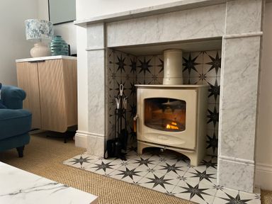A living room corner with a white stove fireplace with black star tile surround a wooden cabinet with a lamp and ceramic vase and a blue armchair at Happy Place in Sheringham