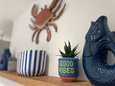 A wooden shelf with a blue fish-shaped pitcher a small plant pot with good vibes text and a blue and white striped pot with a crab decoration on the wall at Happy Place in Sheringham