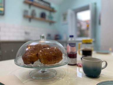 A kitchen table with croissants under a glass dome a mug a jar a bottle and a window in the background at Happy Place in Sheringham