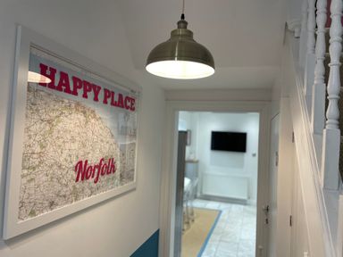 A hallway with a framed map on the wall and a hanging light leading to a room with a wall-mounted TV at Happy Place in Sheringham
