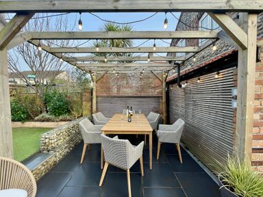 An outdoor dining area with a wooden table and wicker chairs under a pergola with string lights at Happy Place in Sheringham