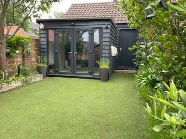 A backyard with artificial grass two potted plants wooden shed with glass doors and a side door surrounded by trees and plants at Happy Place in Sheringham