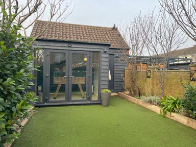 A backyard with artificial grass and plants along the edges with a small building featuring glass double doors at Happy Place in Sheringham
