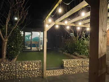 A backyard at night with string lights on a wooden pergola stone walls and a small building with glass doors at Happy Place in Sheringham
