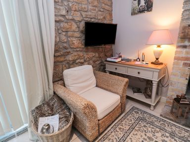 A wicker armchair with cushions next to a basket with a blanket and a dog bowl beside a stone wall a console table with books and a lamp on top and a wall-mounted TV above it at The Coquet Nest in Rothbury