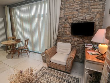 A sitting area with a wicker armchair a small table with two wooden chairs a stone wall with a mounted flat screen tv a lamp on a table and large vertical blinds covering the window at The Coquet Nest in Rothbury