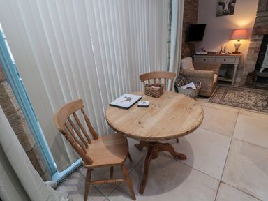 A round wooden table with two wooden chairs beside vertical blinds and a living area with a lamp and armchair at The Coquet Nest in Rothbury