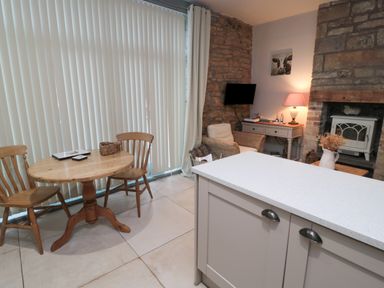 A dining area with wooden table and chairs next to a living space with wicker chair desk lamp and fireplace at The Coquet Nest in Rothbury