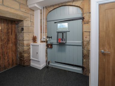 An entryway with a large blue wooden door with metal hinges and latch a small window and a mail basket on it stone walls a small white cabinet with a wooden squirrel figurine on top a wooden door with a metal handle and dark carpeted floor at The Coquet Nest in Rothbury
