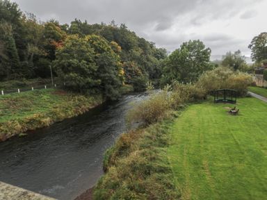 A river flowing alongside a grassy area with benches and trees at The Coquet Nest in Rothbury