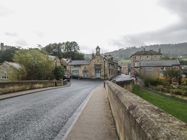 A stone bridge and road leading to stone buildings with parked cars and trees in the background at The Coquet Nest in Rothbury