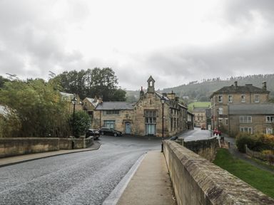 A street with stone buildings and parked cars near a bridge in a village with trees and hills in the background
