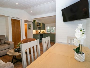 A dining area with white chairs and a wooden table with a plant next to a living room with a sofa and a wooden coffee table and an open kitchen with green cabinets at Esk Pike 9 Skiptory Howe White Cross Bay near Windermere