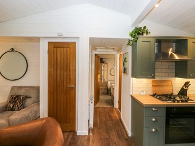 A kitchen with green cabinets and gas stove next to a hallway leading to a bedroom and a living area with a brown chair and gray armchair at Esk Pike 9 Skiptory Howe White Cross Bay near Windermere
