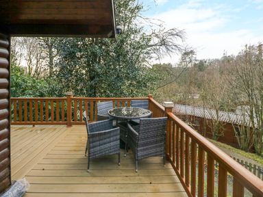 An outdoor wooden deck with a round glass table and four wicker chairs surrounded by trees at Esk Pike 9 Skiptory Howe White Cross Bay near Windermere