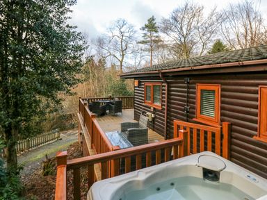 A wooden deck with outdoor seating and a hot tub attached to a log cabin surrounded by trees at Esk Pike 9 Skiptory Howe White Cross Bay near Windermere