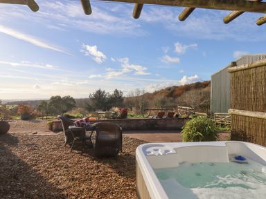 An outdoor patio area with a hot tub and wicker chairs overlooking a garden and trees at Sherway Dairy in Clyst Hydon near Bradninch